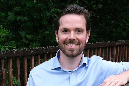 Adam Harley in a blue shirt standing with arm on top of a wooden fence of a bridge..