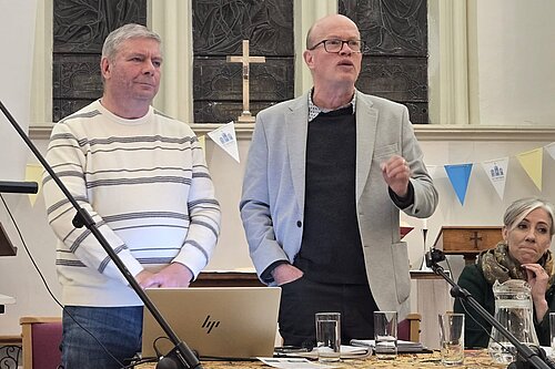 Councillor Tony Lillico, councillor Paul de Kort, and Daisy Cooper MP (from left to right) are pictured during the public meeting. Tony and Paul are both standing, and Paul is speaking to the room. Daisy is sat next to Paul.  
