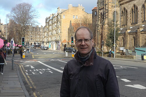 Neil Ross standing at Holy Corner with churches, tenements and blue skies in the background