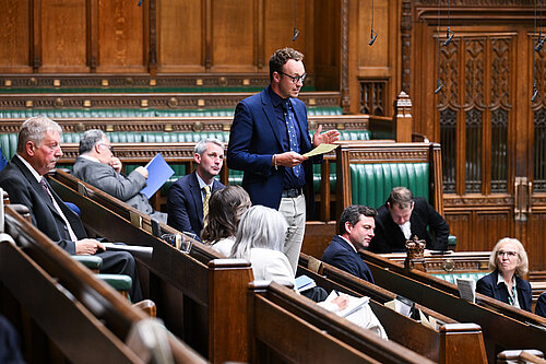A man stands speaking in the House of Commons chamber while other MPs sit and listen.